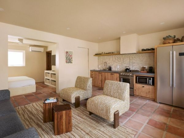 A modern living area with a kitchen, two patterned chairs, a sofa, wooden coffee tables, and a view into a bedroom with a bed and window.