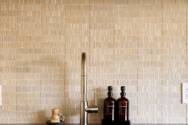 A kitchen sink with a modern faucet, beige tile backsplash and a countertop with two brown bottles and a brush in a dish.