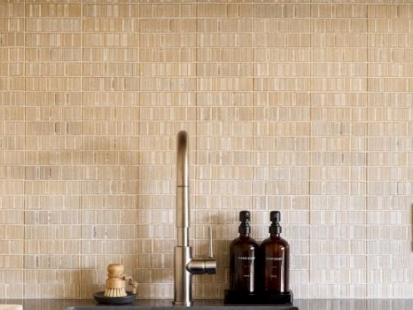 A kitchen sink with a modern faucet, beige tile backsplash and a countertop with two brown bottles and a brush in a dish.
