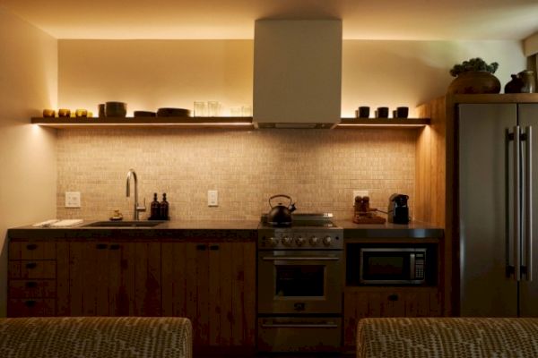 A modern kitchen with ambient lighting, featuring a stove, microwave, refrigerator, and various kitchen items on the countertop and shelf above.