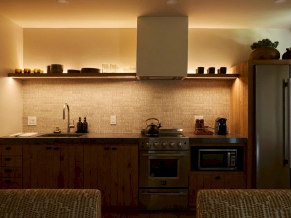 A modern kitchen with ambient lighting, featuring a stove, microwave, refrigerator, and various kitchen items on the countertop and shelf above.