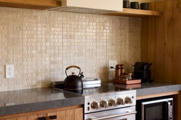 A stylish kitchen with wooden cabinetry, a stovetop with kettle, backsplash tiles, microwave, espresso machine, and various kitchen items.