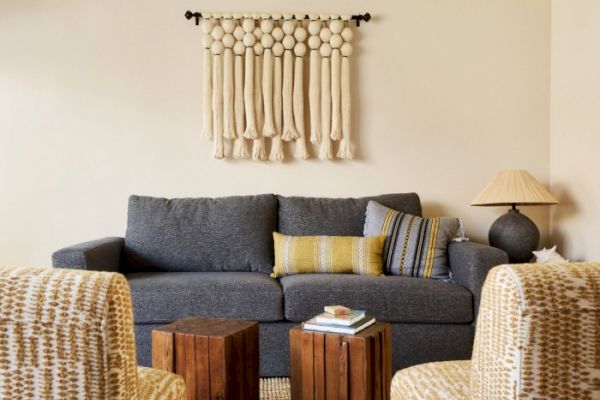 The image shows a cozy living room with a dark gray sofa, two wooden coffee tables, patterned chairs, and a wall hanging above the sofa.