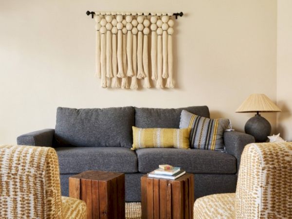 The image shows a cozy living room with a dark gray sofa, two wooden coffee tables, patterned chairs, and a wall hanging above the sofa.