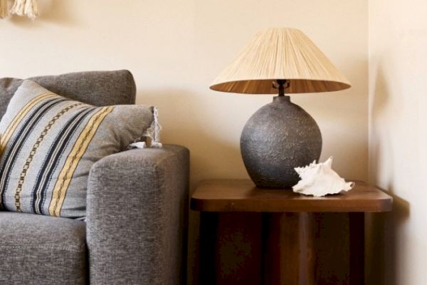 A gray sofa with a striped pillow, a wooden side table holding a textured lamp with a beige shade, and a white conch shell are shown in the image.