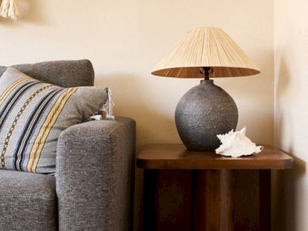 A gray sofa with a striped pillow, a wooden side table holding a textured lamp with a beige shade, and a white conch shell are shown in the image.