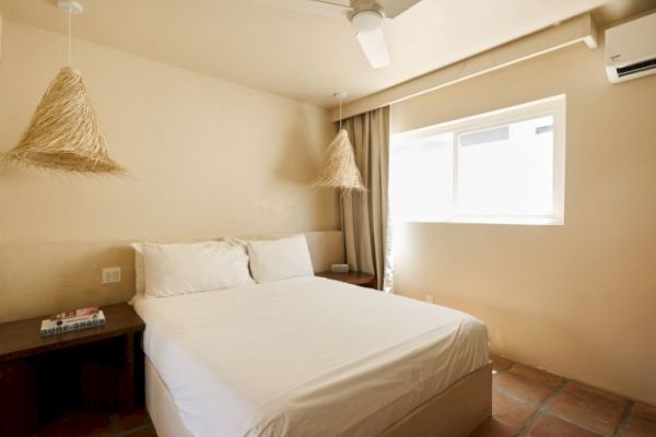 A minimalist bedroom with a double bed, hanging pendant lights, a wooden nightstand with books, a ceiling fan, and a window with curtains.