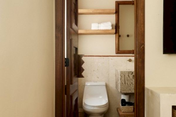 The image shows a small, tidy bathroom with a toilet, a mirror, shelves with towels, and a stone sink underneath.