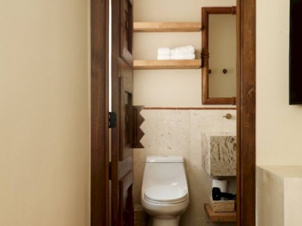 The image shows a small, tidy bathroom with a toilet, a mirror, shelves with towels, and a stone sink underneath.