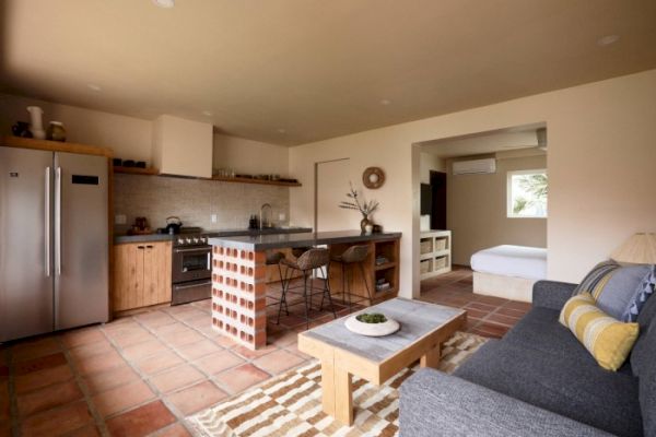 A modern open-plan living kitchen area with a couch, coffee table, island, stainless steel appliances, and adjacent bedroom visible in the background.