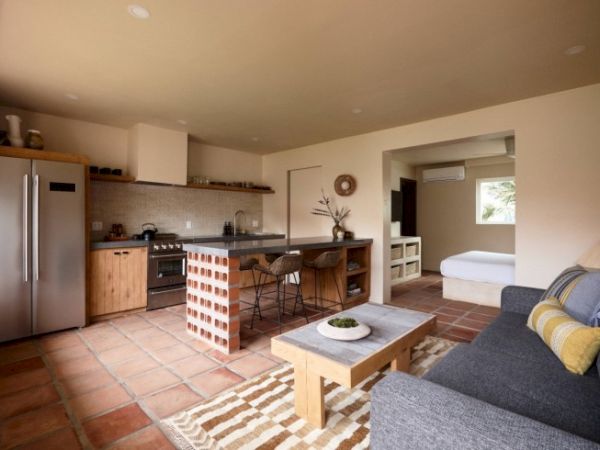 A modern open-plan living kitchen area with a couch, coffee table, island, stainless steel appliances, and adjacent bedroom visible in the background.