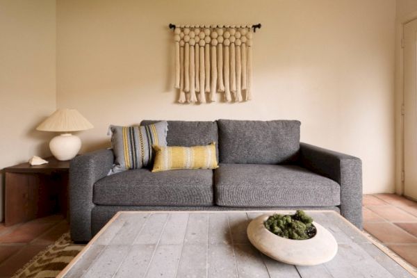 A cozy living room with a gray sofa, decorative pillows, a wall hanging, a wooden coffee table with a centerpiece, and a side table with a lamp.