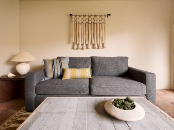 A cozy living room with a gray sofa, decorative pillows, a wall hanging, a wooden coffee table with a centerpiece, and a side table with a lamp.