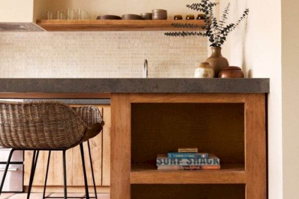 A cozy kitchen features wooden shelves, a gray countertop, wicker stools, various pottery, and a cookbook titled 