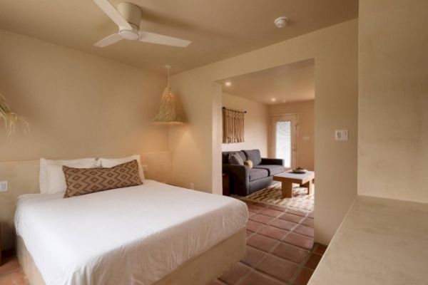 The image shows a cozy bedroom with a double bed, a ceiling fan, and a connected living area with a sofa and coffee table, featuring warm, earthy tones.