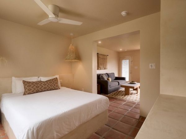 The image shows a cozy bedroom with a double bed, a ceiling fan, and a connected living area with a sofa and coffee table, featuring warm, earthy tones.