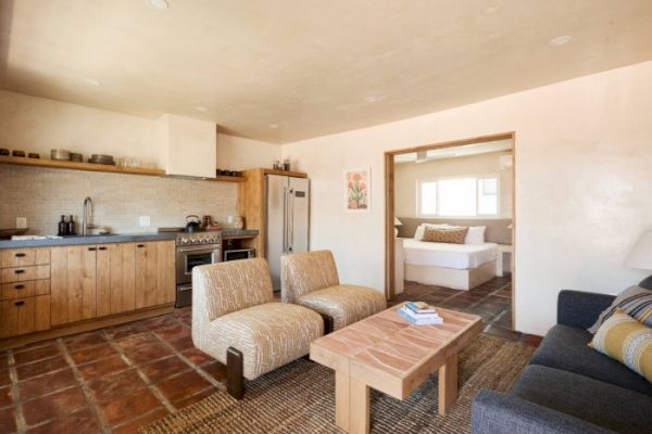 A cozy living area with kitchen, two chairs, a coffee table, and a couch. An opening shows a bedroom with a bed and window at the far end.