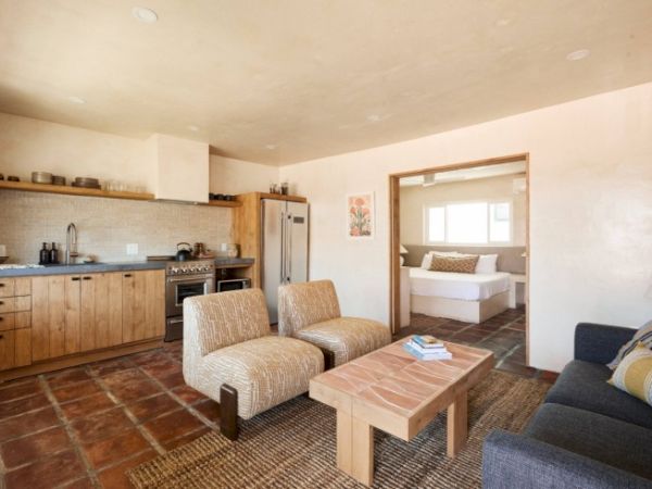 A cozy living area with kitchen, two chairs, a coffee table, and a couch. An opening shows a bedroom with a bed and window at the far end.