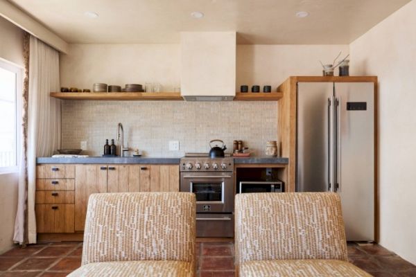 A modern kitchen with wooden cabinets, a stainless steel fridge, stove, and sink, with open shelving above and two patterned chairs in the foreground.