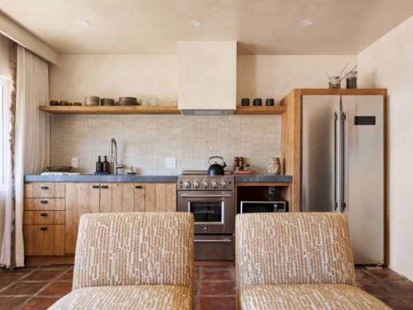 A modern kitchen with wooden cabinets, a stainless steel fridge, stove, and sink, with open shelving above and two patterned chairs in the foreground.