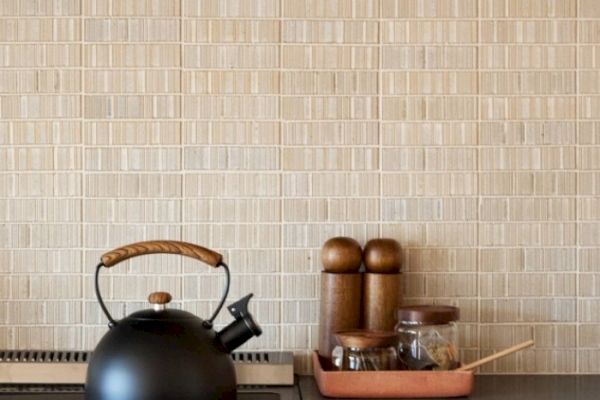 A black kettle with a wooden handle sits on a stove. Nearby are containers for spices and utensils arranged neatly in a tray, all against a tiled wall.