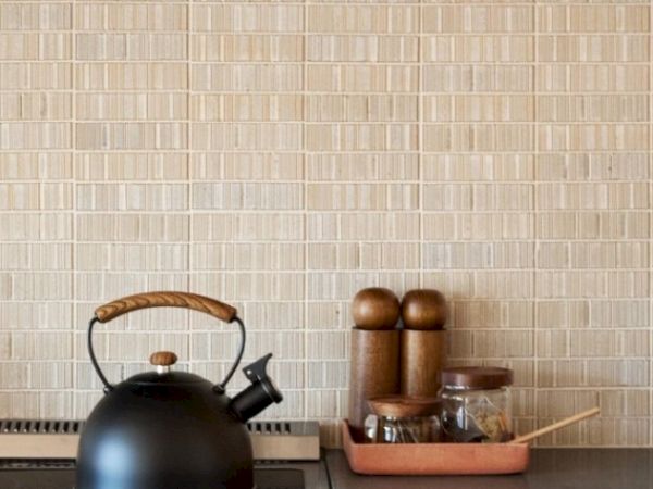 A black kettle with a wooden handle sits on a stove. Nearby are containers for spices and utensils arranged neatly in a tray, all against a tiled wall.