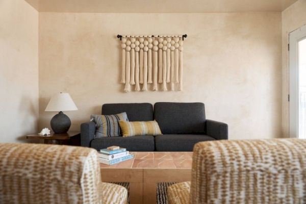 A cozy living room with a dark sofa, patterned armchairs, a lamp on a side table, books on a central table, and wall decor above the sofa.