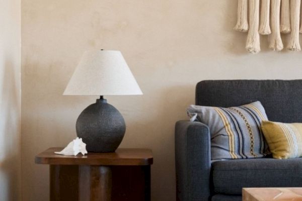 The image shows a cozy corner with a grey sofa topped with colorful cushions, a wooden side table with a lamp, and a decorative shell.