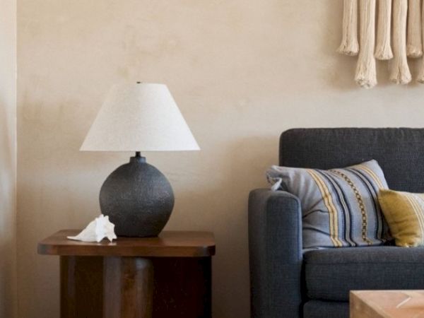 The image shows a cozy corner with a grey sofa topped with colorful cushions, a wooden side table with a lamp, and a decorative shell.