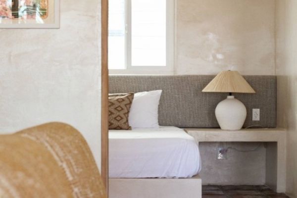 The image shows a cozy bedroom corner with a white bed, a grey headboard, a patterned pillow, and a bedside table with a lamp next to a window.