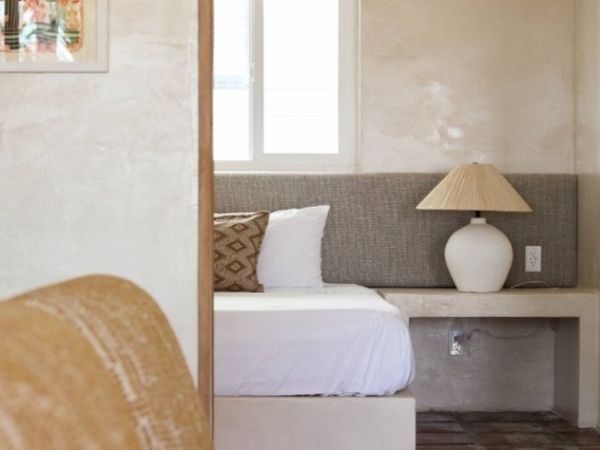 The image shows a cozy bedroom corner with a white bed, a grey headboard, a patterned pillow, and a bedside table with a lamp next to a window.
