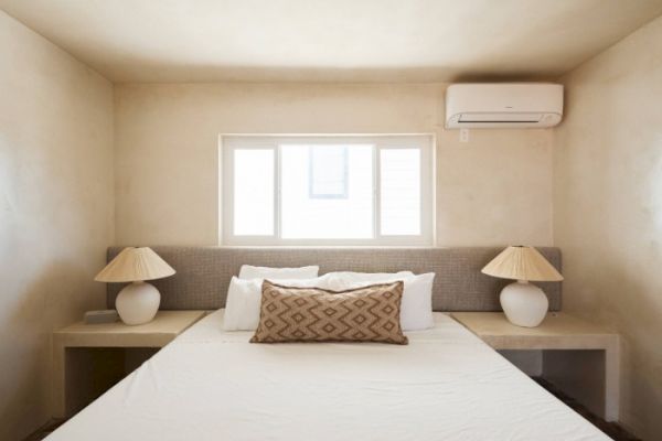 A minimalist bedroom features a bed with neutral bedding, two bedside lamps, a window, and an air conditioner on the wall.