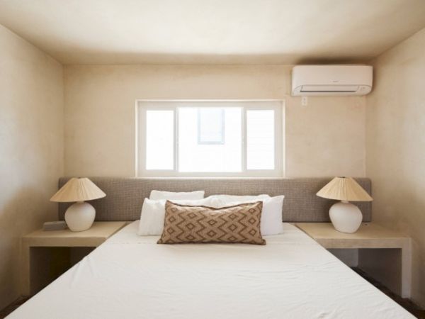 A minimalist bedroom features a bed with neutral bedding, two bedside lamps, a window, and an air conditioner on the wall.