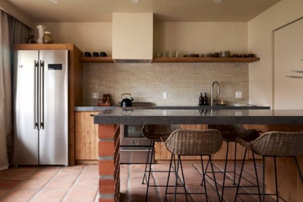 A modern kitchen features a stainless steel fridge, wooden cabinets, an island with barstools, and neutral decor with tiled backsplash and flooring.