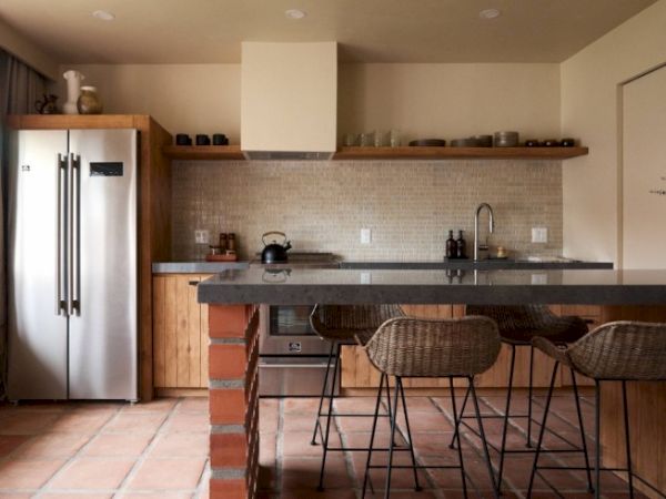 A modern kitchen features a stainless steel fridge, wooden cabinets, an island with barstools, and neutral decor with tiled backsplash and flooring.