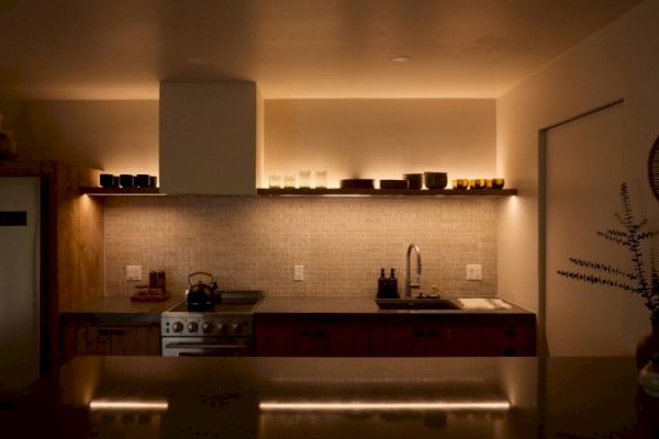 A warmly lit modern kitchen with minimalist decor, featuring a stove, sink, shelves, and various kitchen items on countertops and shelves.