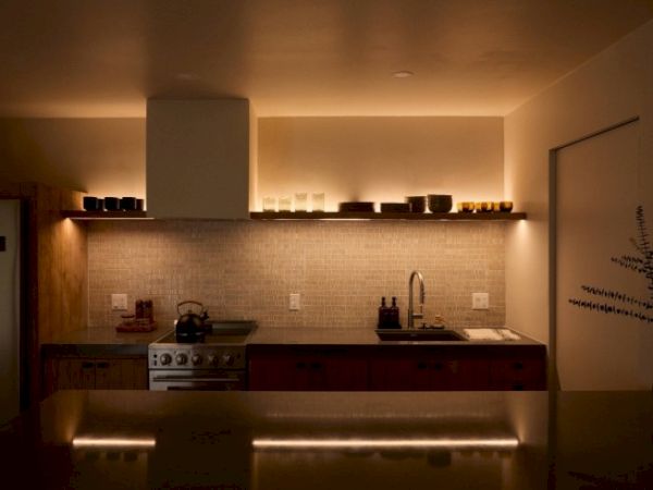 A warmly lit modern kitchen with minimalist decor, featuring a stove, sink, shelves, and various kitchen items on countertops and shelves.