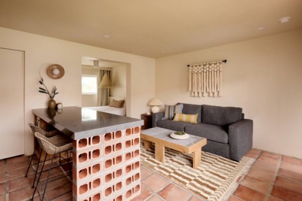 A cozy living space featuring a gray sofa, wooden coffee table, open brick kitchen counter with bar stools, wall decor, and a patterned rug on tiled floor.