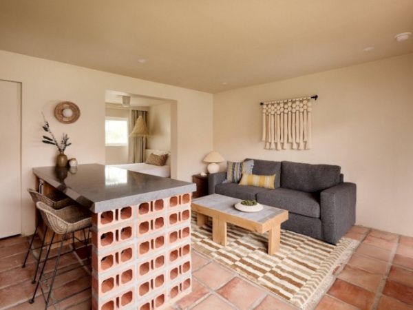 A cozy living space featuring a gray sofa, wooden coffee table, open brick kitchen counter with bar stools, wall decor, and a patterned rug on tiled floor.