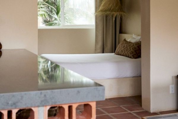 The image shows a cozy room with a bed, pillows, and a window, featuring a tiled floor and a countertop with visible brickwork under it.