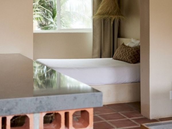 The image shows a cozy room with a bed, pillows, and a window, featuring a tiled floor and a countertop with visible brickwork under it.