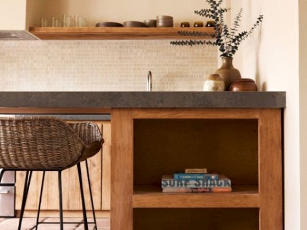 A modern kitchen with wooden shelves, a woven chair, vases on the countertop, and books on the lower shelf, creating a cozy atmosphere.