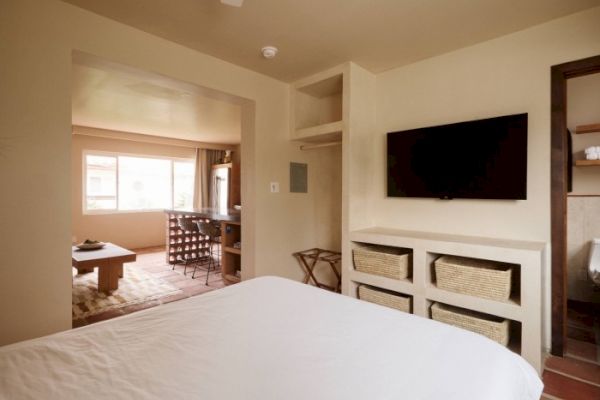 A cozy hotel room features a bed, a wall-mounted TV, storage baskets, and an open view into a sitting area with a table and chairs near a large window.