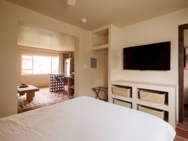 A cozy hotel room features a bed, a wall-mounted TV, storage baskets, and an open view into a sitting area with a table and chairs near a large window.