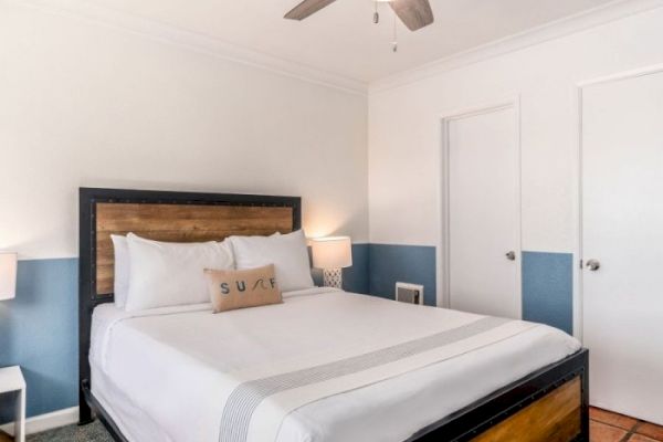 A neatly arranged bedroom with a bed, white linens, two bedside tables with lamps, a ceiling fan, and two doors. The wall is half-painted blue.