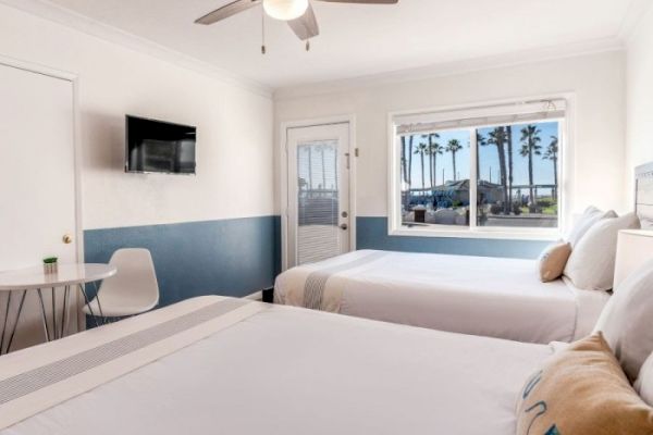 This image shows a hotel room with two beds, a table with a white chair, a wall-mounted TV, a ceiling fan, and a window with a view of palm trees.