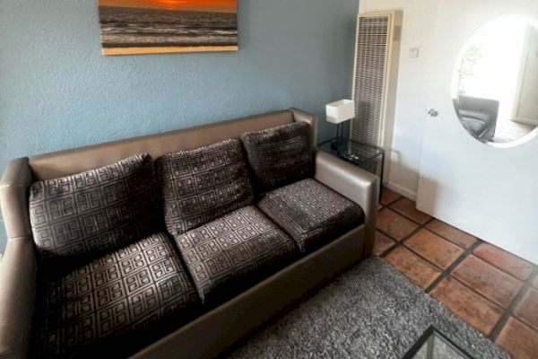 A living room with a patterned sofa, a beach sunset picture, a side table, lamp, heating unit, and a door with an oval mirror.