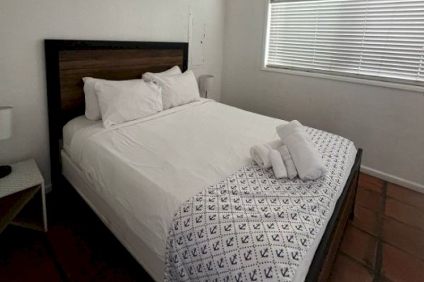 A neatly made bed with white linens and pillows, a patterned blanket at the foot, and rolled towels sits in a minimalistic room with blinds on the window.