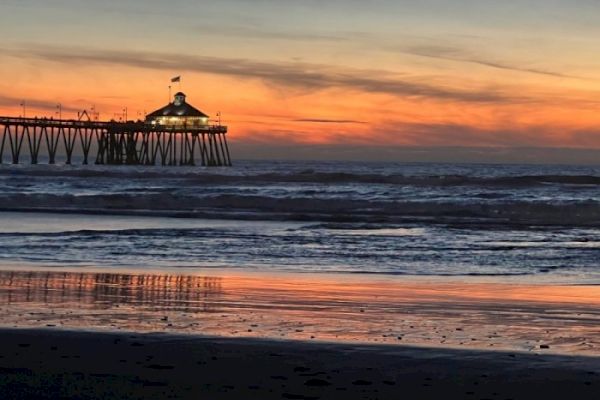 A tranquil beach scene at sunset with an illuminated pier and ocean waves reflecting the vibrant colors of the sky.