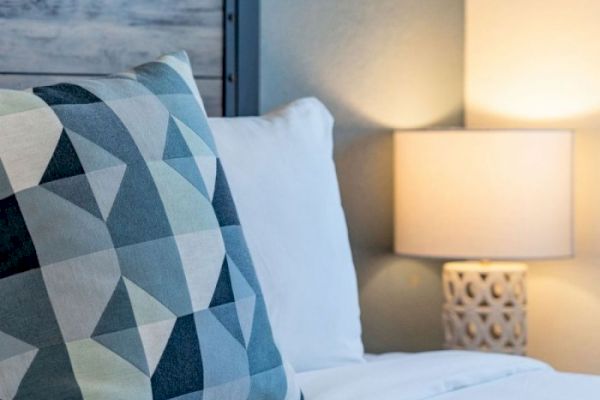 The image features a bed with white pillows and a geometric-patterned throw pillow, next to a lit bedside lamp with a decorative base.